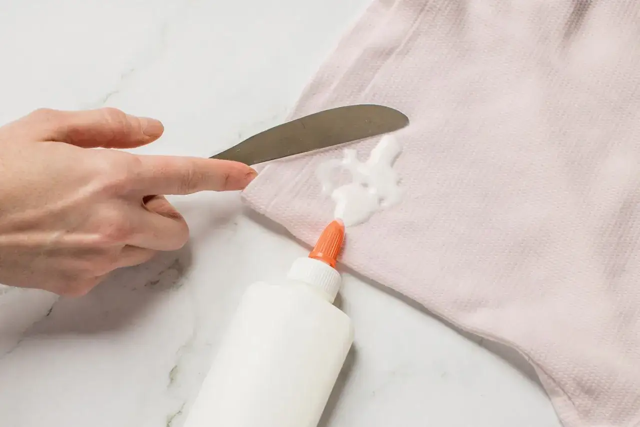 Maintenant, comment enlever de la colle n&eacute;opr&egrave;ne sur du tissu ? Une main utilise un couteau pour gratter une tache de colle sur un tissu rose p&acirc;le.