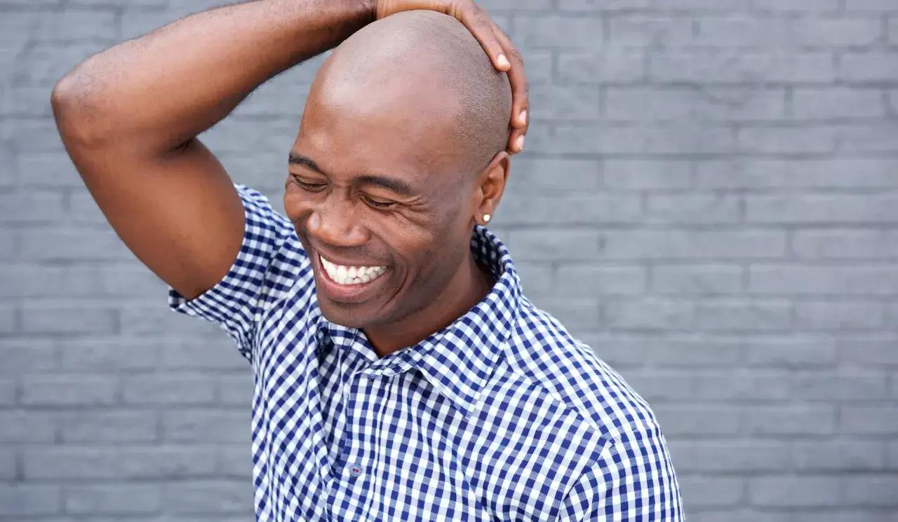 Hombre sonriente se toca la cabeza reci&eacute;n afeitada. Viste una camisa a cuadros azules y blancas.