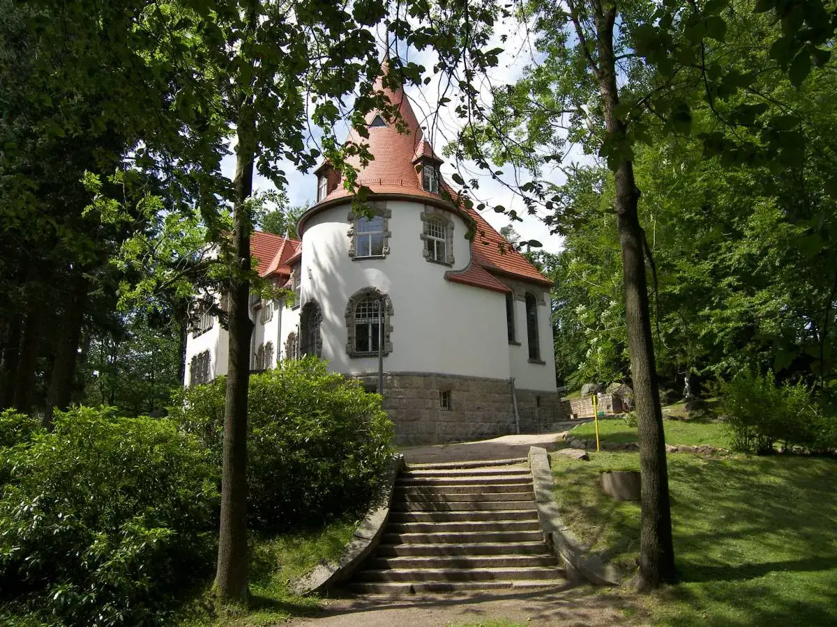 Biały dom z czerwoną wieżą, otoczony zielenią. Schody prowadzą do wejścia. Miejsce jak z bajki o jagniątków.