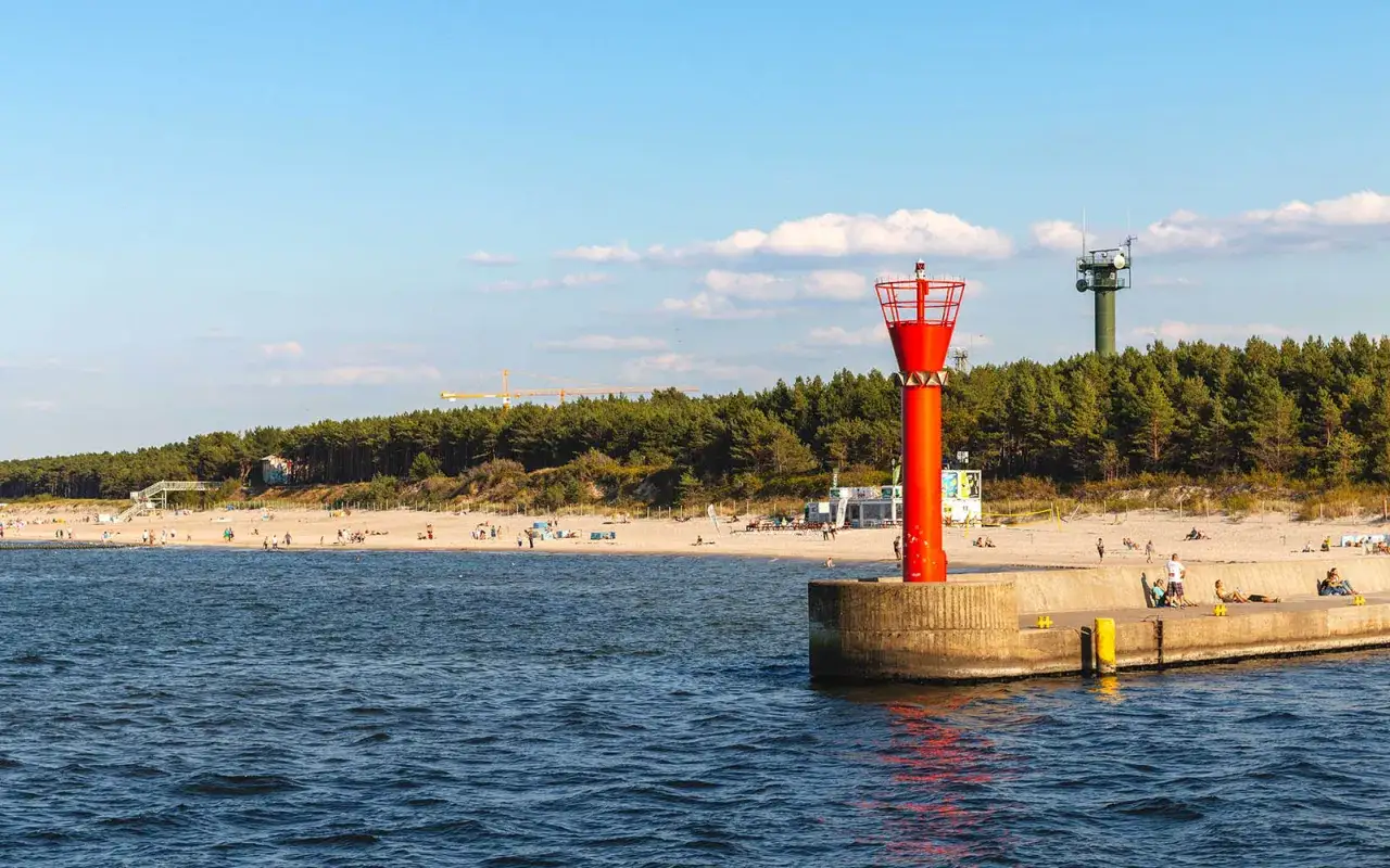 Mrzeżyno atrakcje: czerwona i zielona wieża nawigacyjna na tle plaży i lasu. Ludzie odpoczywają na molo i piasku.