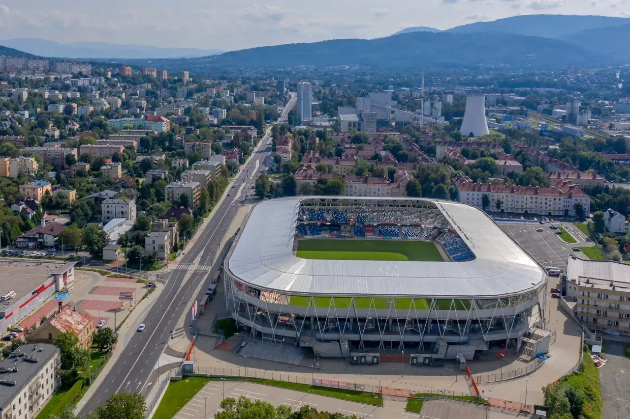 Nowoczesny stadion Podbeskidzie z zieloną murawą i trybunami w Biesku-Białej, otoczony miejską zabudową i górami.