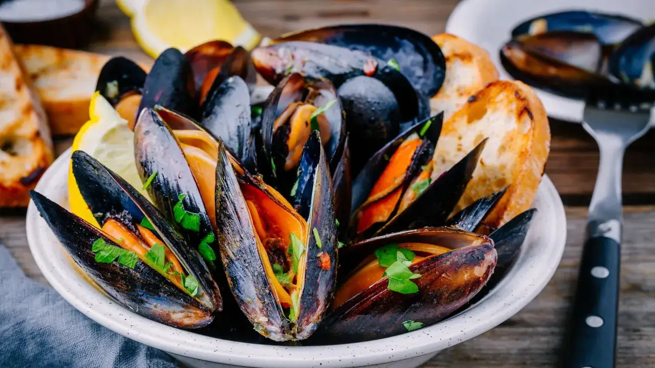 Mejillones en salsa, servidos en un cuenco blanco con pan tostado y rodajas de limón.
