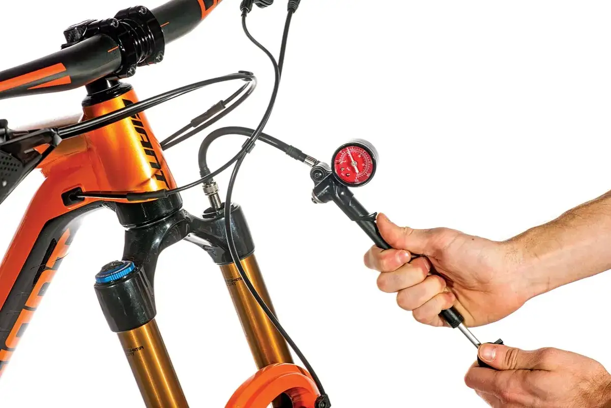 Adjusting the mtb suspension setup with a shock pump. A hand holds the pump, connected to the fork, with a pressure gauge visible.