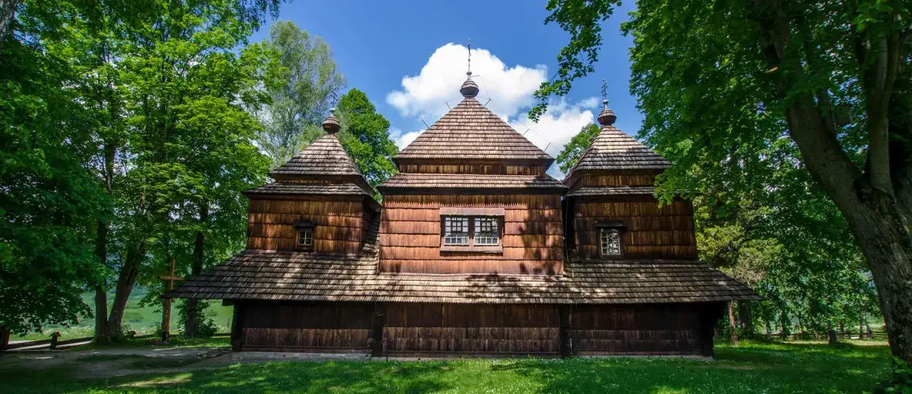 Drewniana cerkiew z charakterystyczną czarną górną częścią, otoczona zielenią drzew.