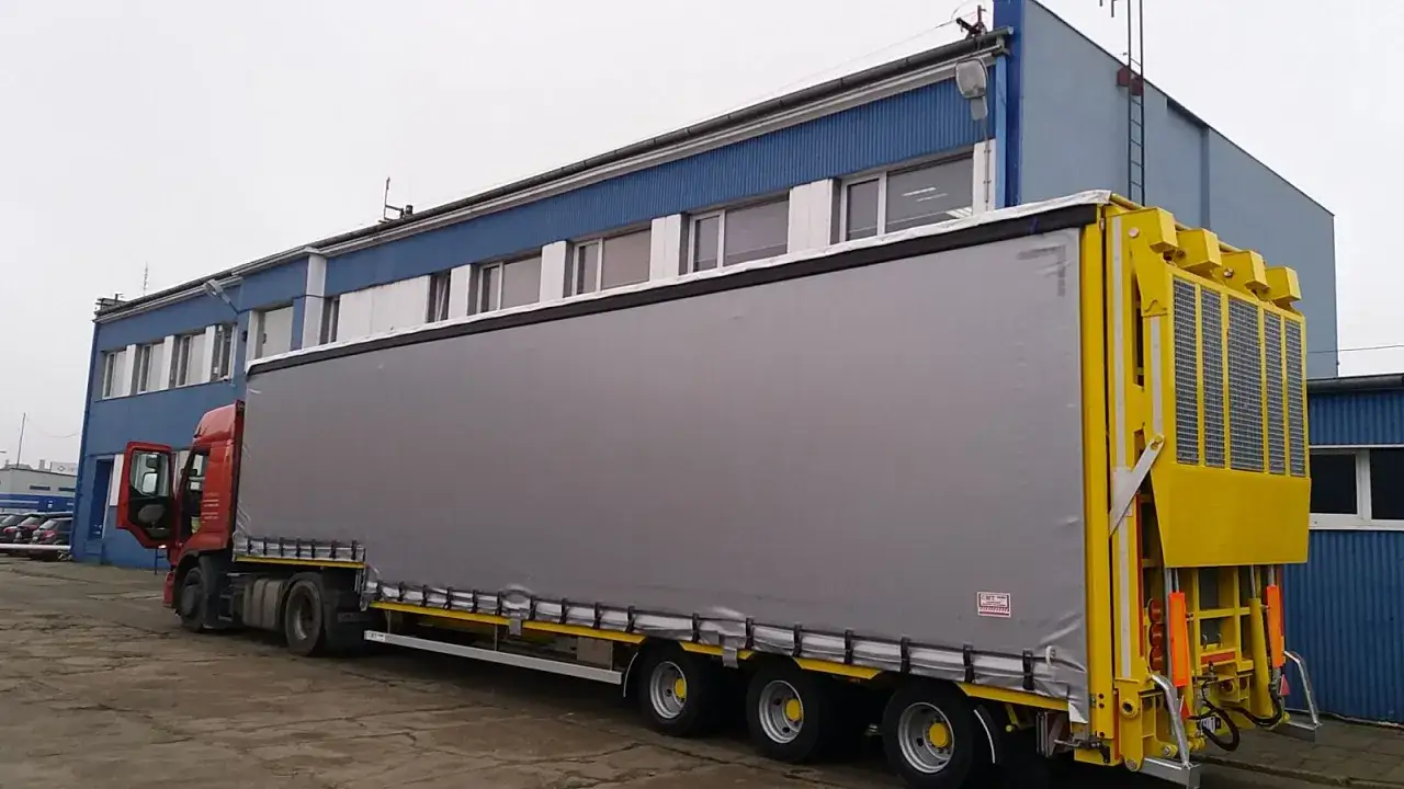 Czerwona ciężarówka z naczepą jumbo stoi przed niebieskim budynkiem. Naczepa ma szare plandeki i żółty tył.