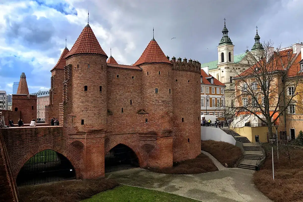 Warszawska Barbakana, symbol Starego Miasta, z ceglanymi wieżami i dachami. W tle kościół i kamienice.