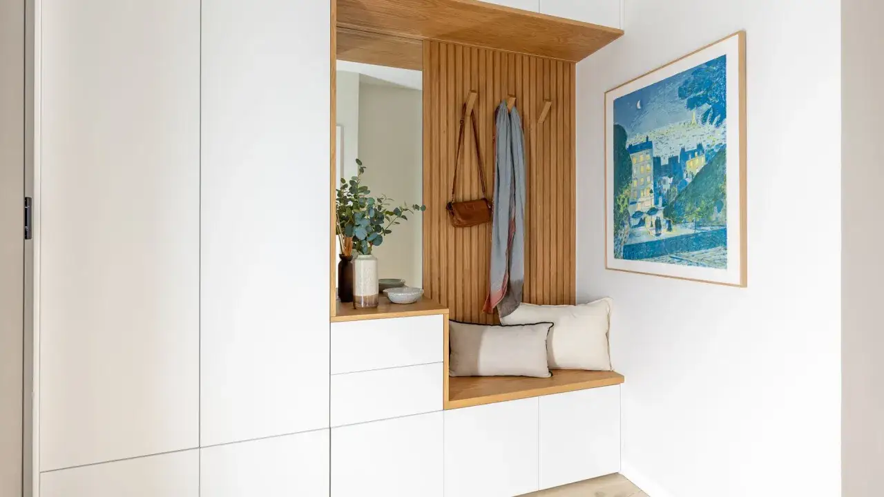 Un recibidor moderno con banco de madera, espejo, perchero con bolso y bufanda, y un cuadro de paisaje urbano. Un espacio acogedor con entradas pequeñas con encanto.