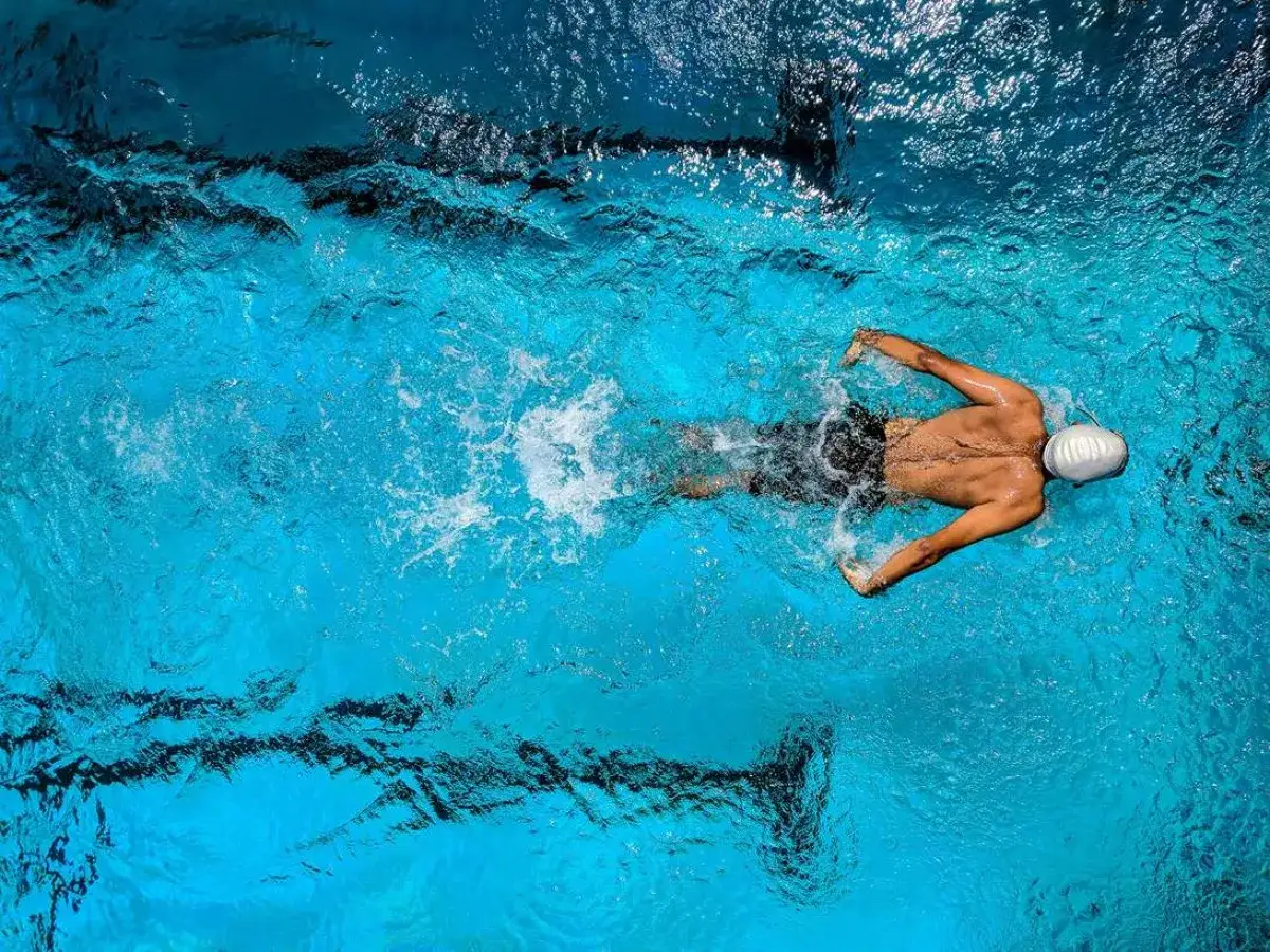 A swimmer glides through blue water, demonstrating how to swim for exercise. The overhead shot captures the powerful strokes and the wake left behind.