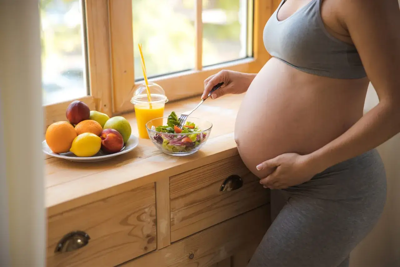 Gravid kvinna med illamående äter sallad vid fönstret. Frukt och juice står på träbänken.