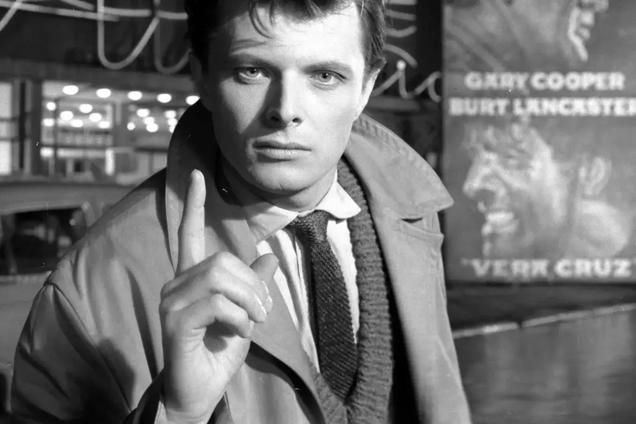 Młody Andrzej Maj, z palcem uniesionym w geście ostrzeżenia, stoi przed plakatem filmu 