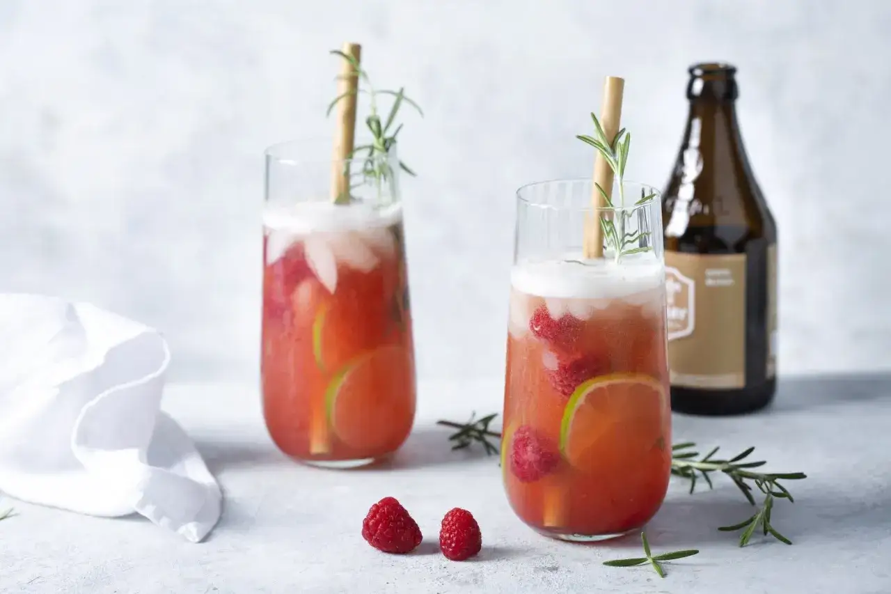 Deux verres de cocktails fruités, garnies de framboises et de rondelles de citron vert, avec une touche de romarin. Une bouteille de bière est visible en arrière-plan.