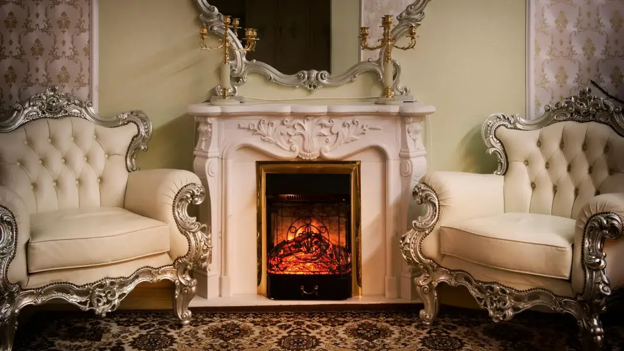 A cozy fireplace glows in this opulent Victorian interior design, flanked by plush, cream-colored armchairs with ornate silver detailing.