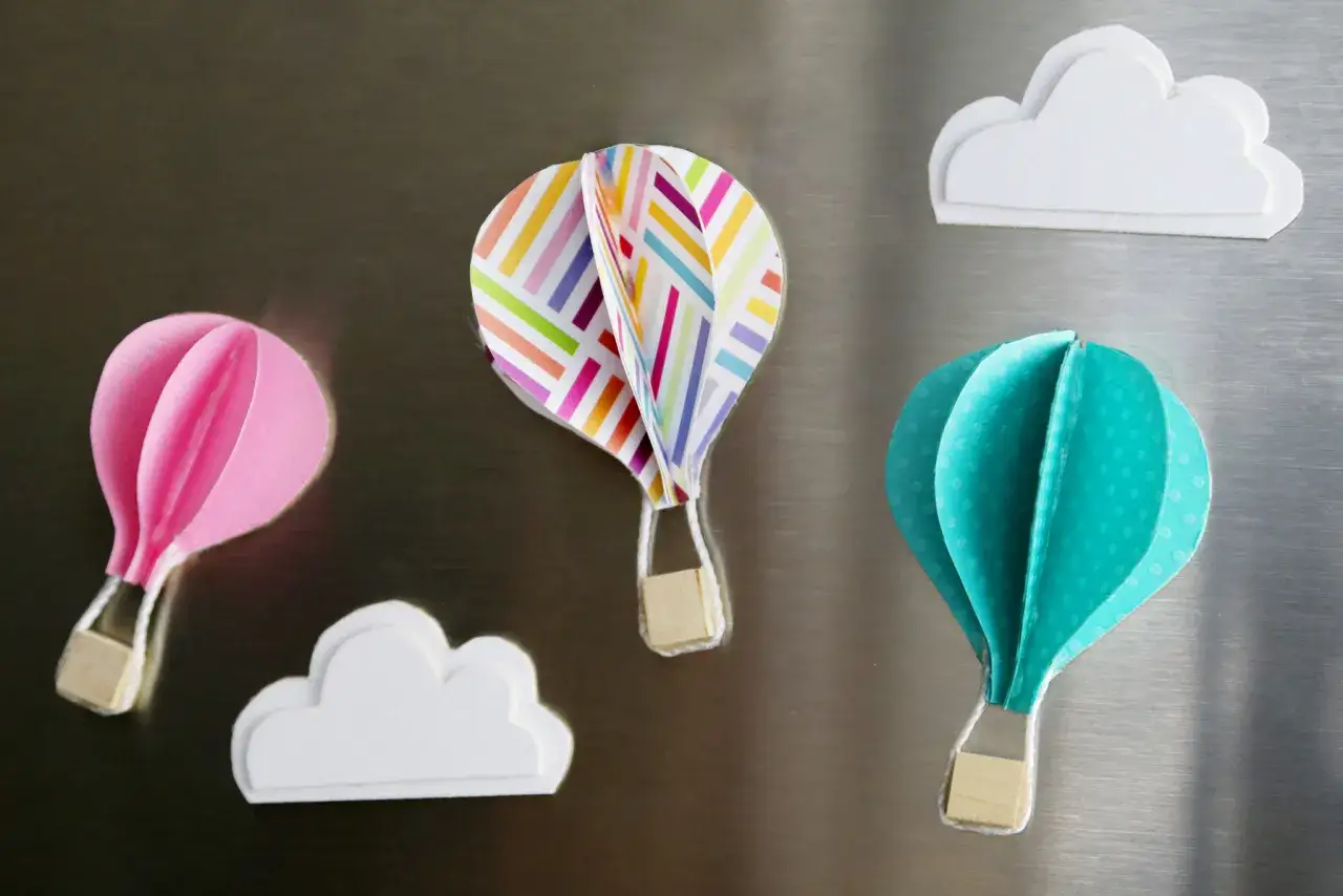 Globos aerostáticos de manualidades, uno rosa, uno multicolor y uno turquesa, flotan junto a nubes blancas sobre una superficie metálica.