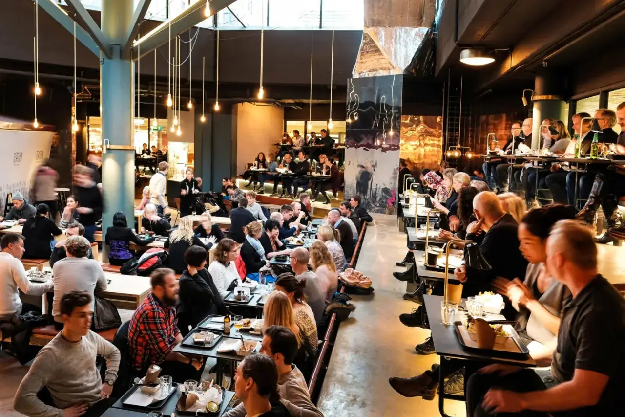 Folk äter och umgås på en livlig **Ringen food court**. Många bord är upptagna, stämningen är social och maten ser god ut.