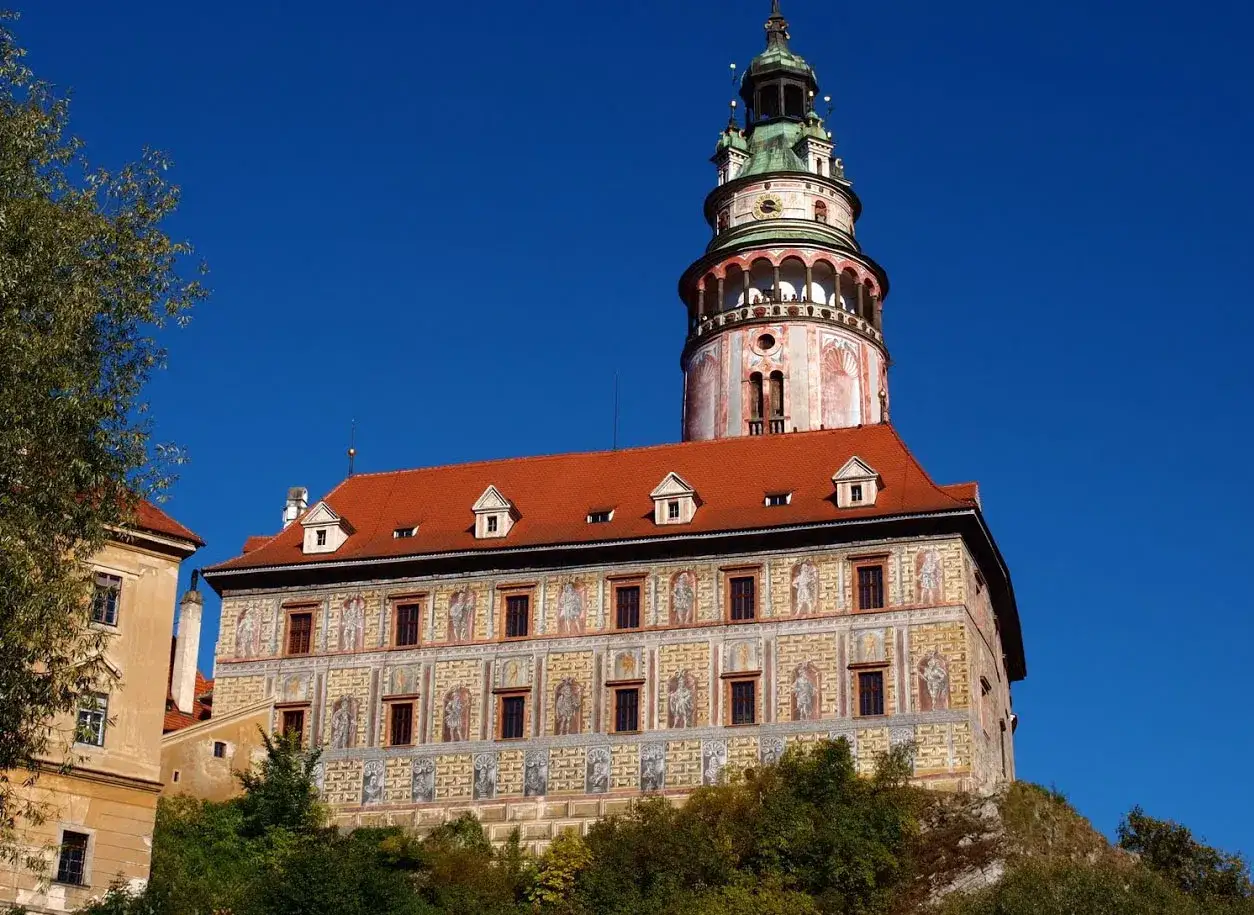 Czeski Krumlov zamek, z charakterystyczną wieżą i zdobioną fasadą, wznosi się na wzgórzu pod błękitnym niebem.