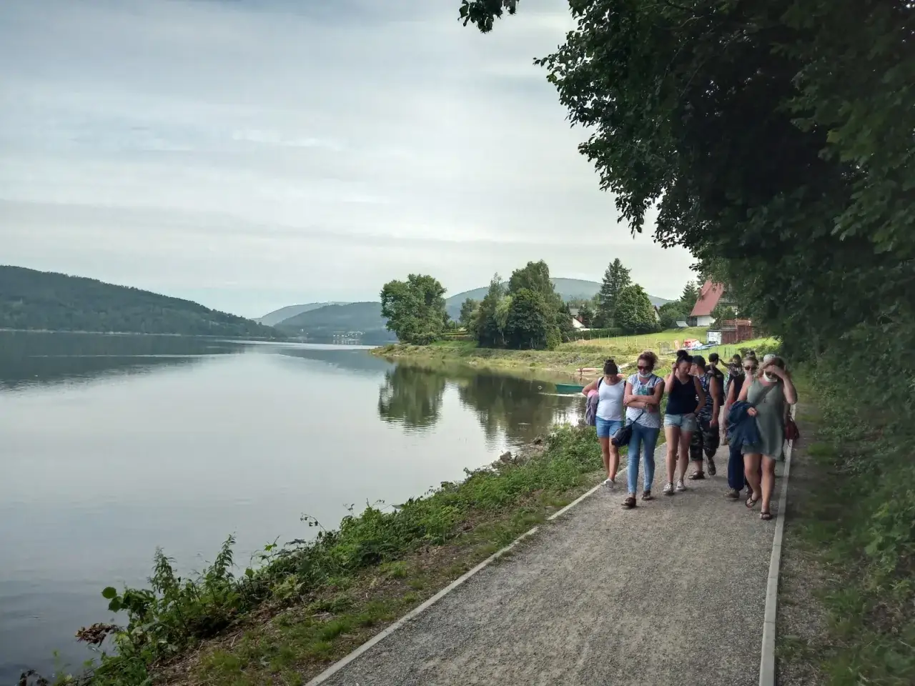 Grupa os&oacute;b spaceruje ścieżką nad spokojnym jeziorem żywieckim, otoczonym zielonymi wzg&oacute;rzami.
