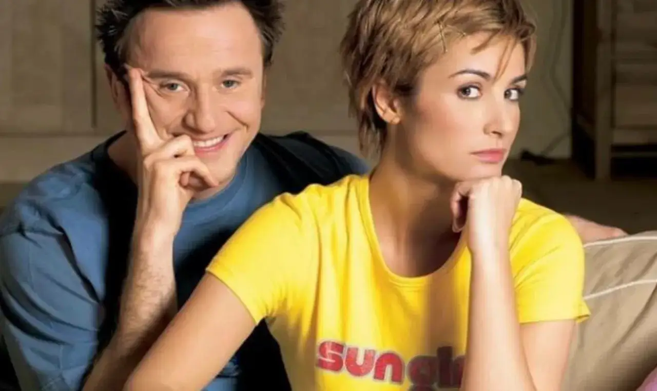 Joanna Brodzik i mężczyzna w niebieskiej koszulce, uśmiechnięty, opiera głowę na dłoni. Ona w żółtej bluzce, zamyślona.
