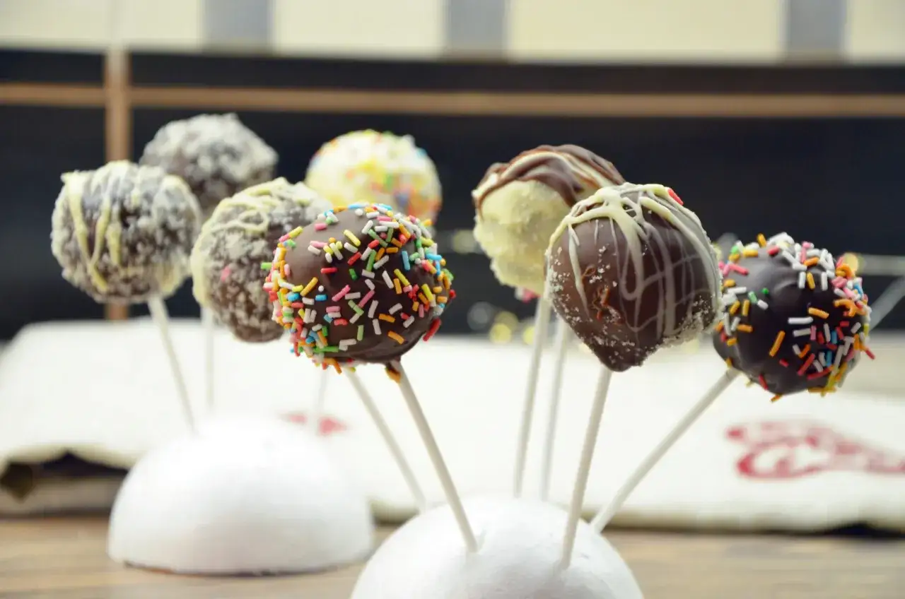 Kolorowe cake pops z jasnego biszkoptu, oblane czekoladą i posypane kolorową posypką lub wiórkami.