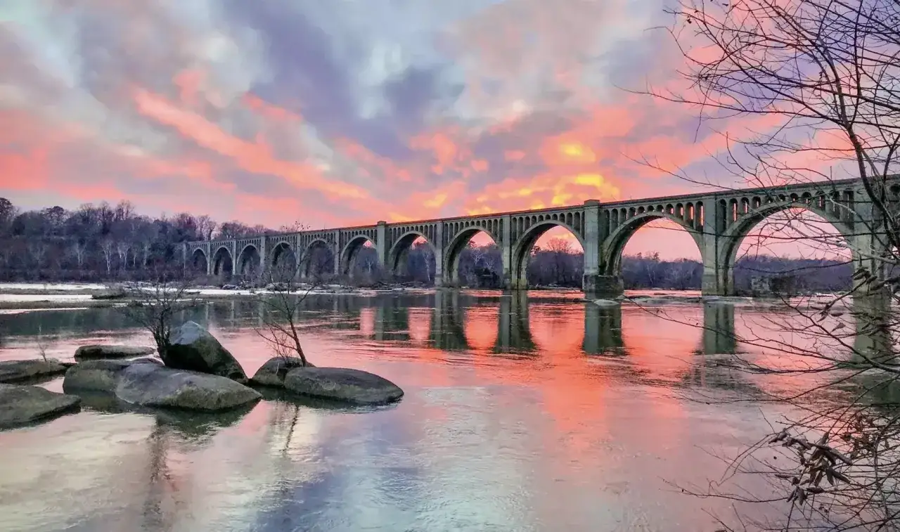 Richmond, Virginia's iconic Mayo Bridge arches over the James River at sunset, reflecting the vibrant sky. A must-see sight!