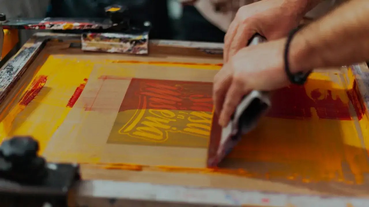 Manos aplicando tinta amarilla y roja sobre un dise&ntilde;o con serigraf&iacute;a que es una t&eacute;cnica de impresi&oacute;n.