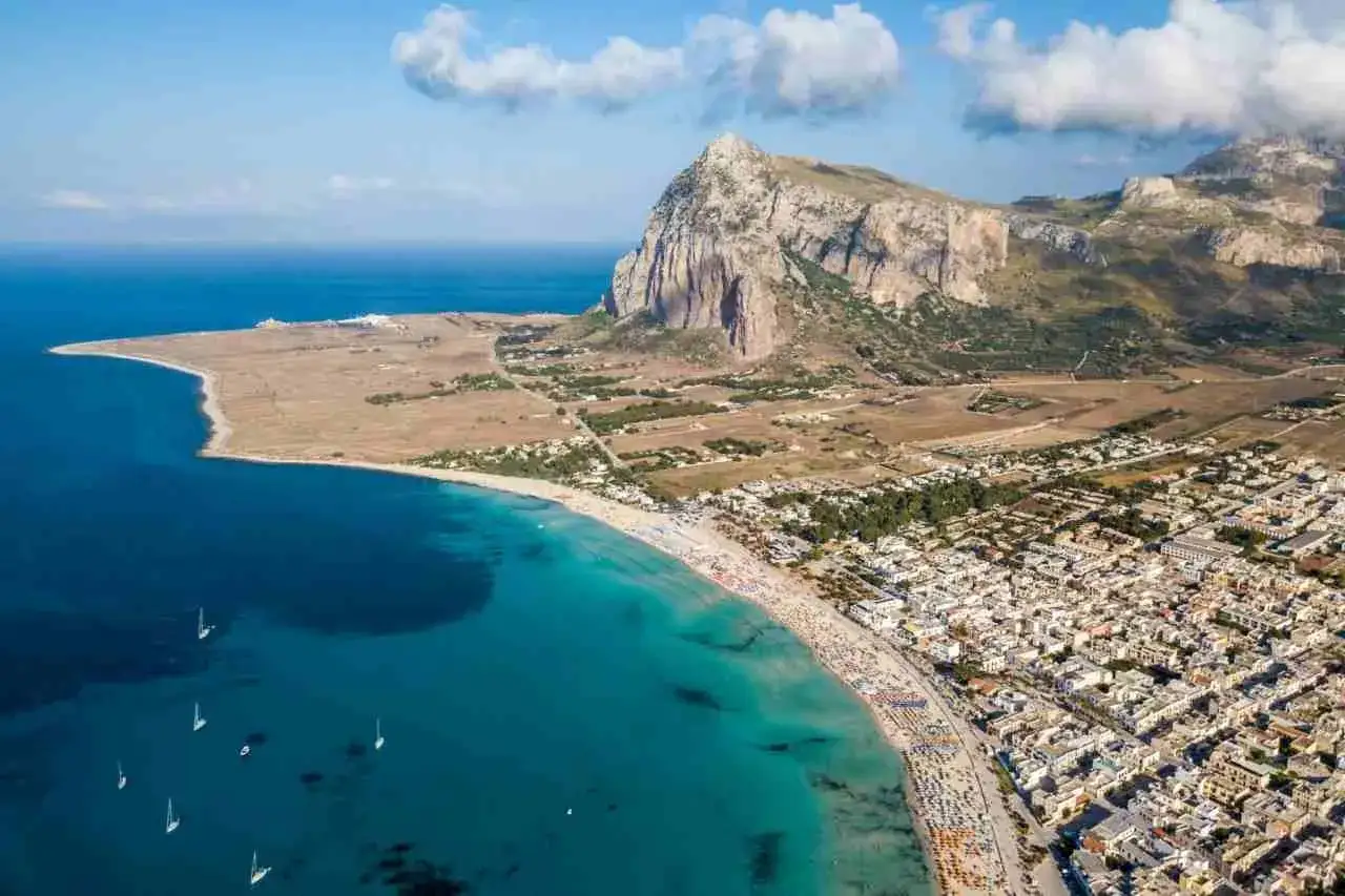 Vista aerea di una spiaggia affollata e del mare cristallino in Sicilia, con un imponente monte sullo sfondo.