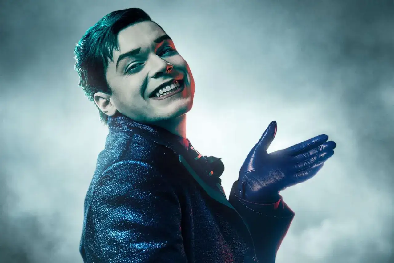 Cameron Monaghan w roli Jokera, znany z filmów i seriali, w błyszczącym garniturze i niebieskich rękawiczkach.