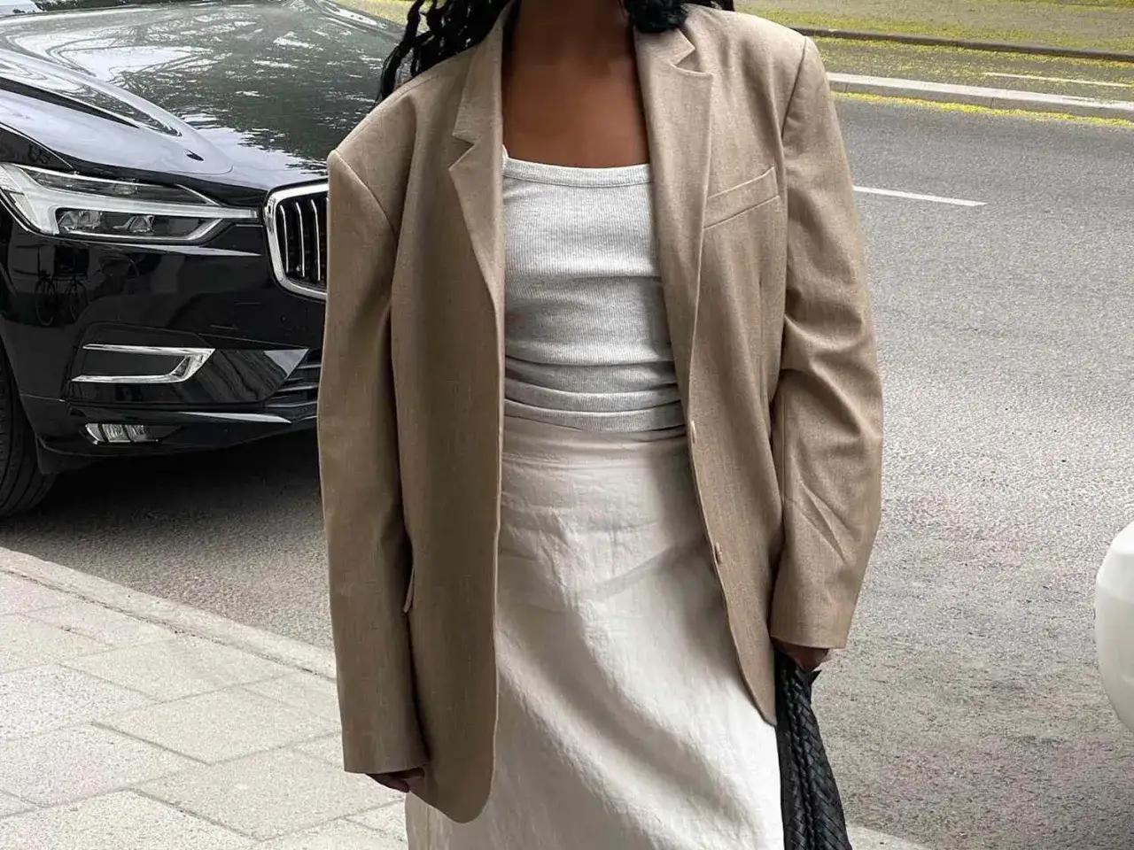 Elegancka, clean girl w beżowej marynarce i białej spódnicy, obok czarnego samochodu.
