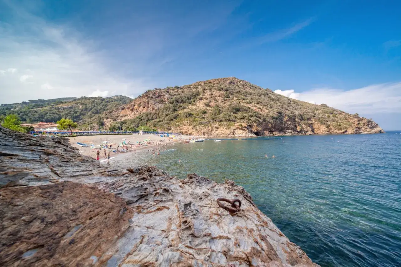 Spiaggia affollata con ombrelloni e gente che nuota, sullo sfondo una collina verdeggiante e il mare cristallino della Toscana, Isola d'Elba.