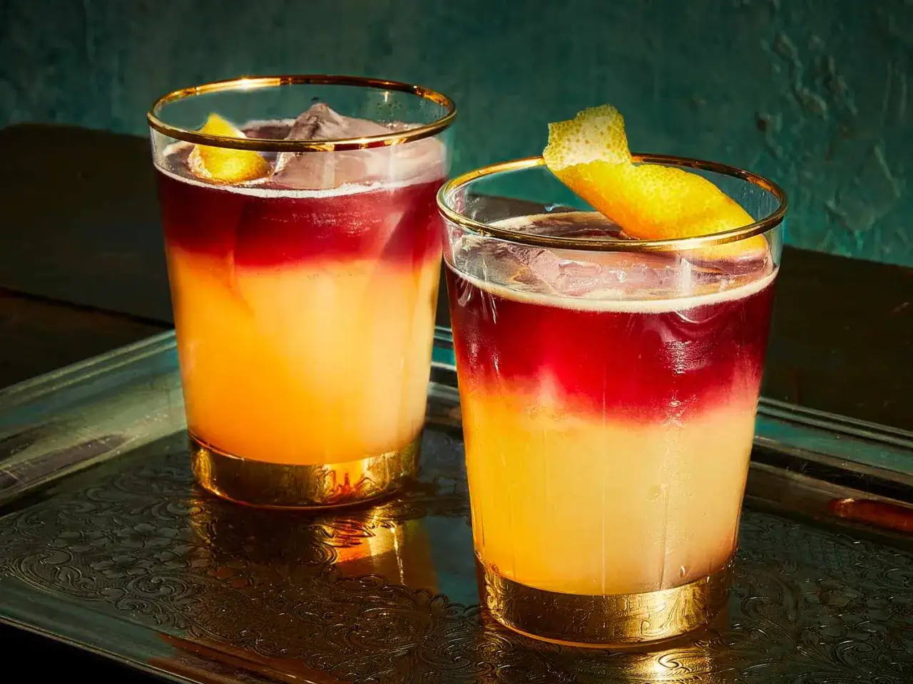 Tv&aring; vackra New York Sours med lager av r&ouml;tt och gult, garnerade med citronskal, serveras i eleganta glas p&aring; en bricka.