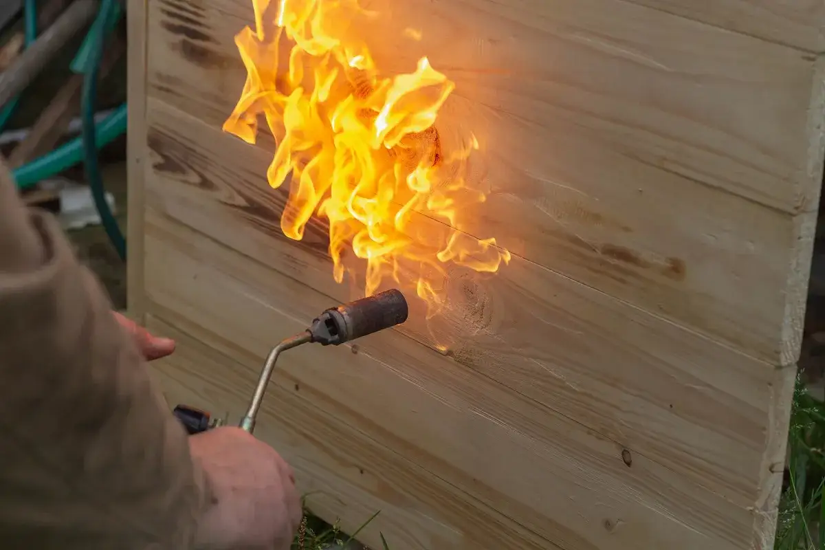 Flammen lodern auf hellem Holz, das sehr alt ist und schlecht brennt. Ein Mann h&auml;lt einen Brenner.