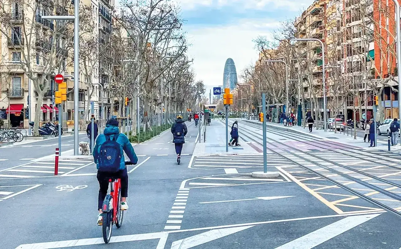 Radfahrer und E-Scooter-Fahrer nutzen die breiten Wege in den **Superblocks Barcelona**. Tramschienen und Bäume säumen die Straße.