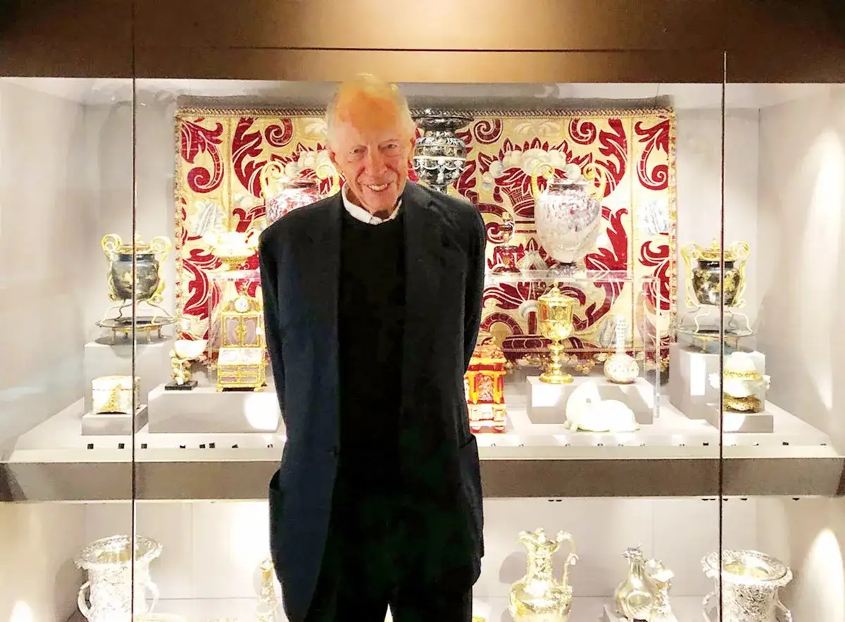 A man smiles in front of a display case filled with ornate vases, golden chalices, and decorative boxes, part of the Rothschild art collection.