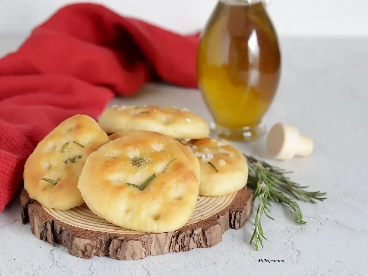 Focaccine di semola dorate, profumate al rosmarino, su un tagliere di legno. Accanto, una bottiglia d'olio e un tovagliolo rosso.