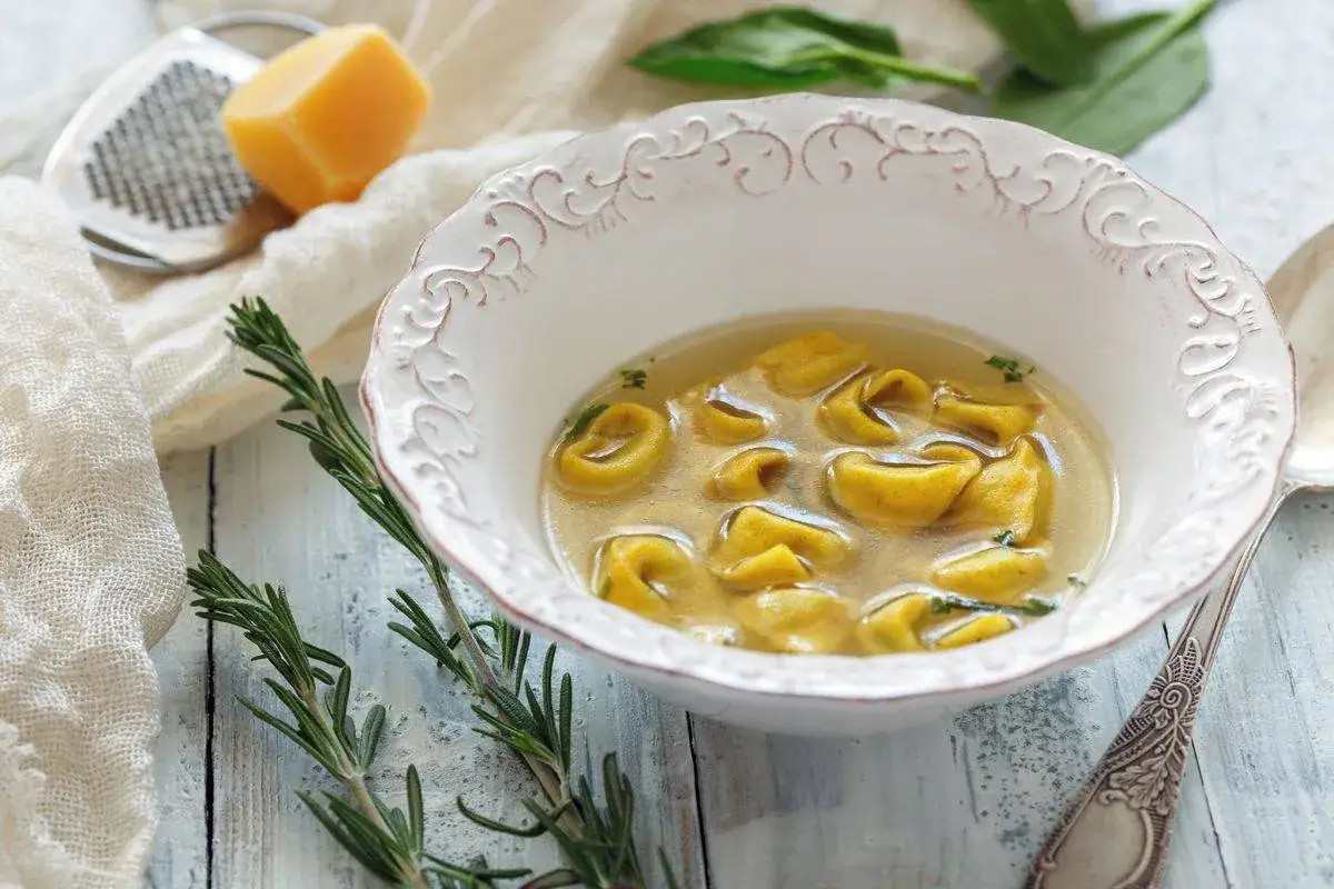 Un piatto invitante di tortellini in brodo, con rosmarino fresco e un pezzo di parmigiano sullo sfondo.