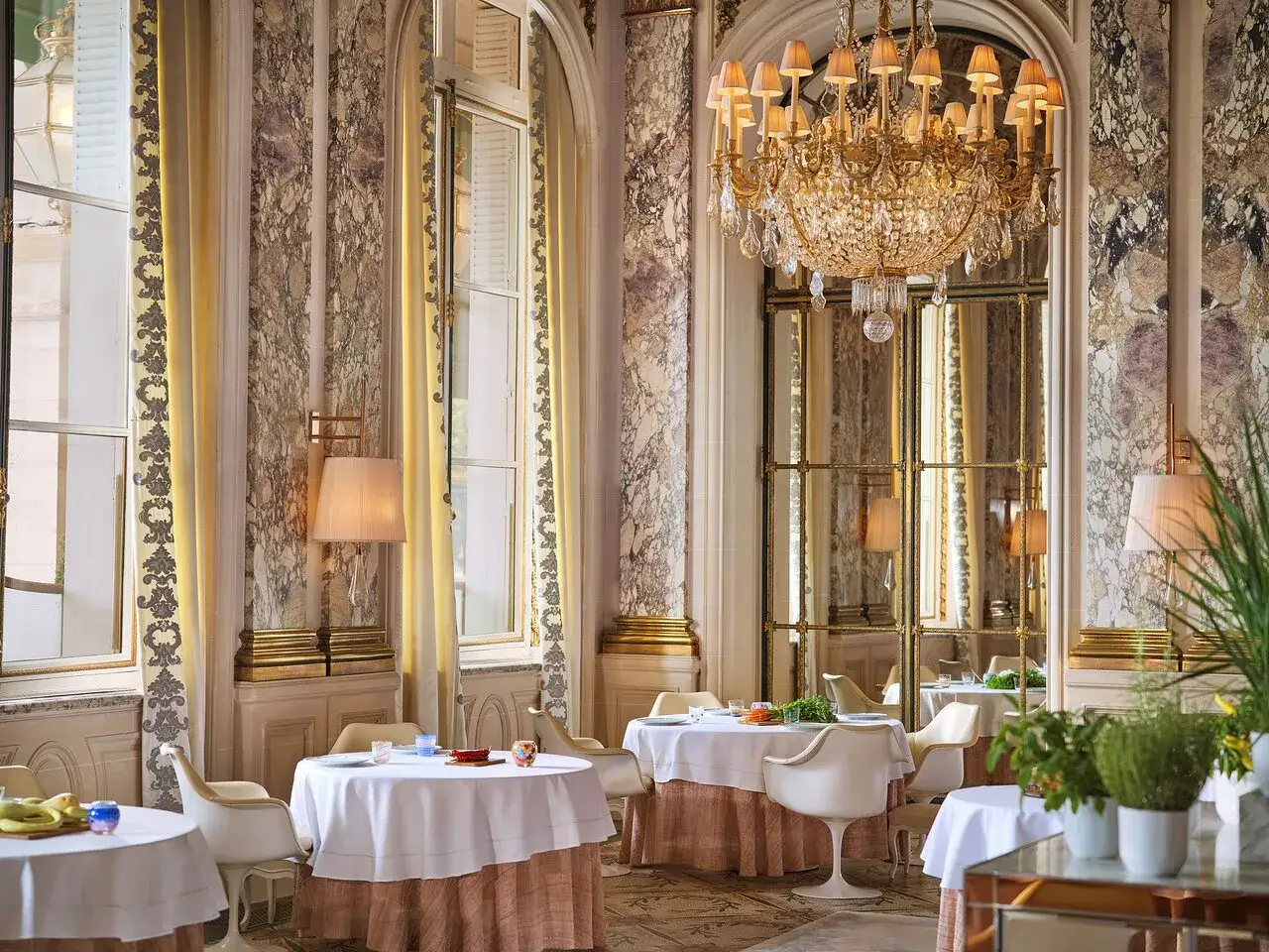 Lussuosa sala da pranzo con tavoli apparecchiati, lampadario dorato e marmi eleganti, un assaggio dell'esperienza culinaria di Alain Ducasse a Parigi.