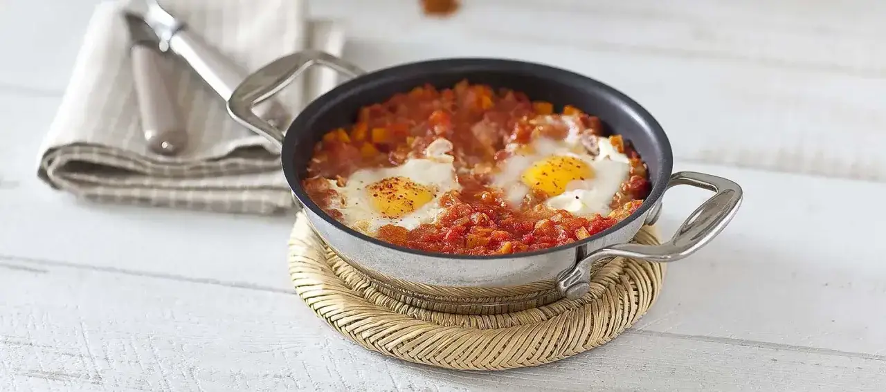 Un plat d'oeuf à la tomate mijoté dans une poêle, servi sur un dessous de plat en osier.