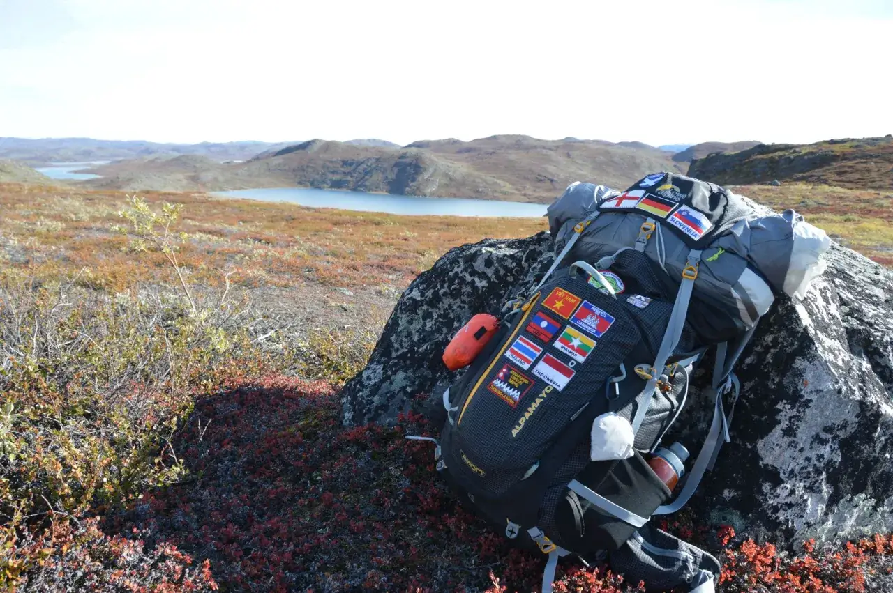 Ein Rucksack eines thru hikers, beladen mit Flaggen-Aufn&auml;hern, ruht auf einem Felsen in einer weiten, h&uuml;geligen Landschaft mit Seen.