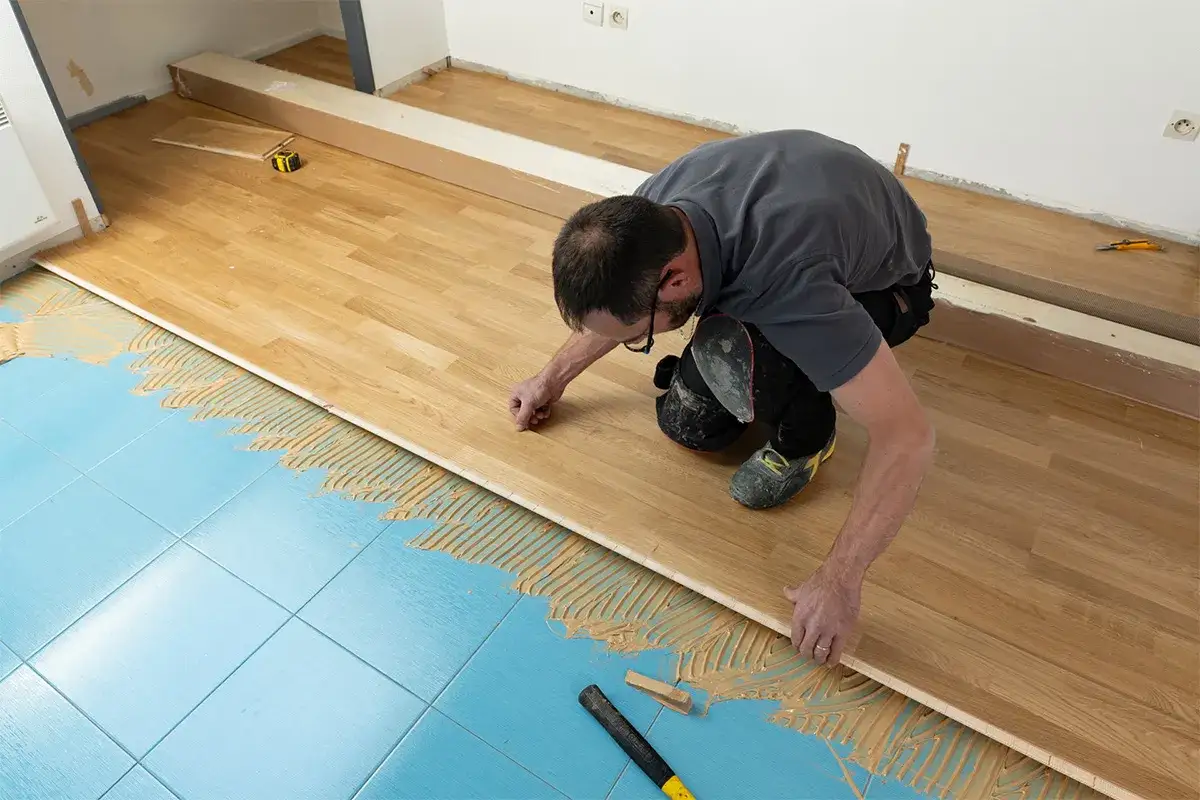 Pose d'un parquet stratifié sur un sol bleu carrelé, avec de la colle appliquée. L'homme ajuste une lame de bois.