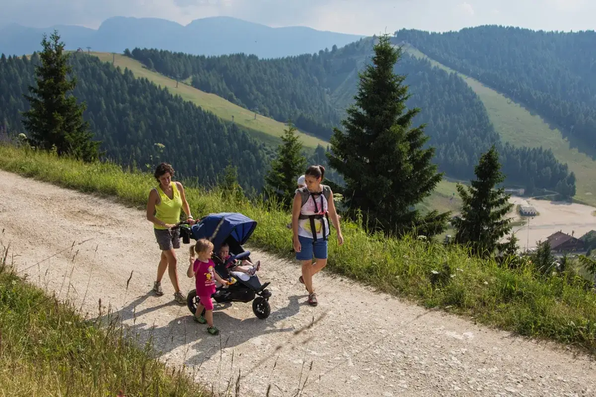 Due donne passeggiano con un passeggino e un bimbo in braccio su un sentiero di montagna. I neonati godono dell'aria fresca e del panorama.