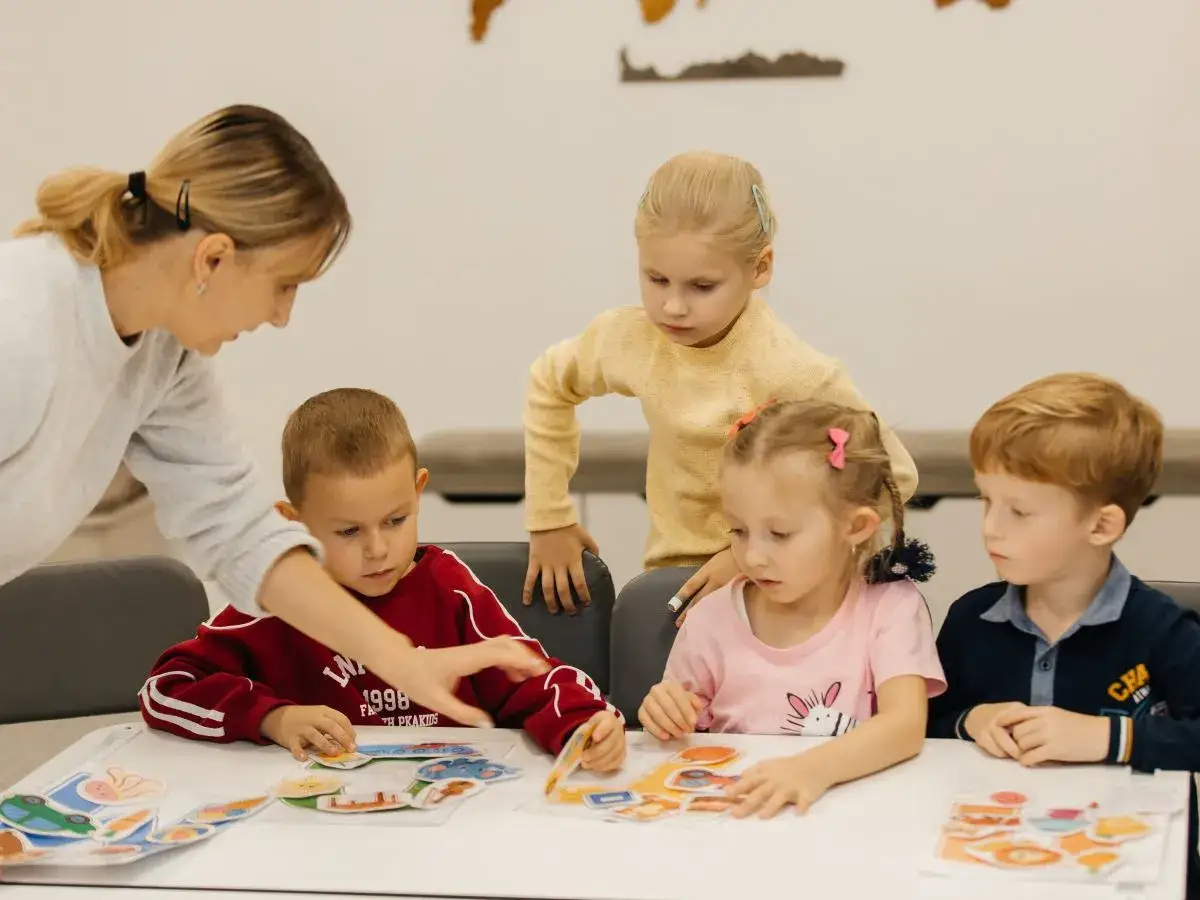 Nauczycielka pomaga chłopcu z niepokojącymi zachowaniami 5-latka układać puzzle. Dzieci skupione na zabawie.