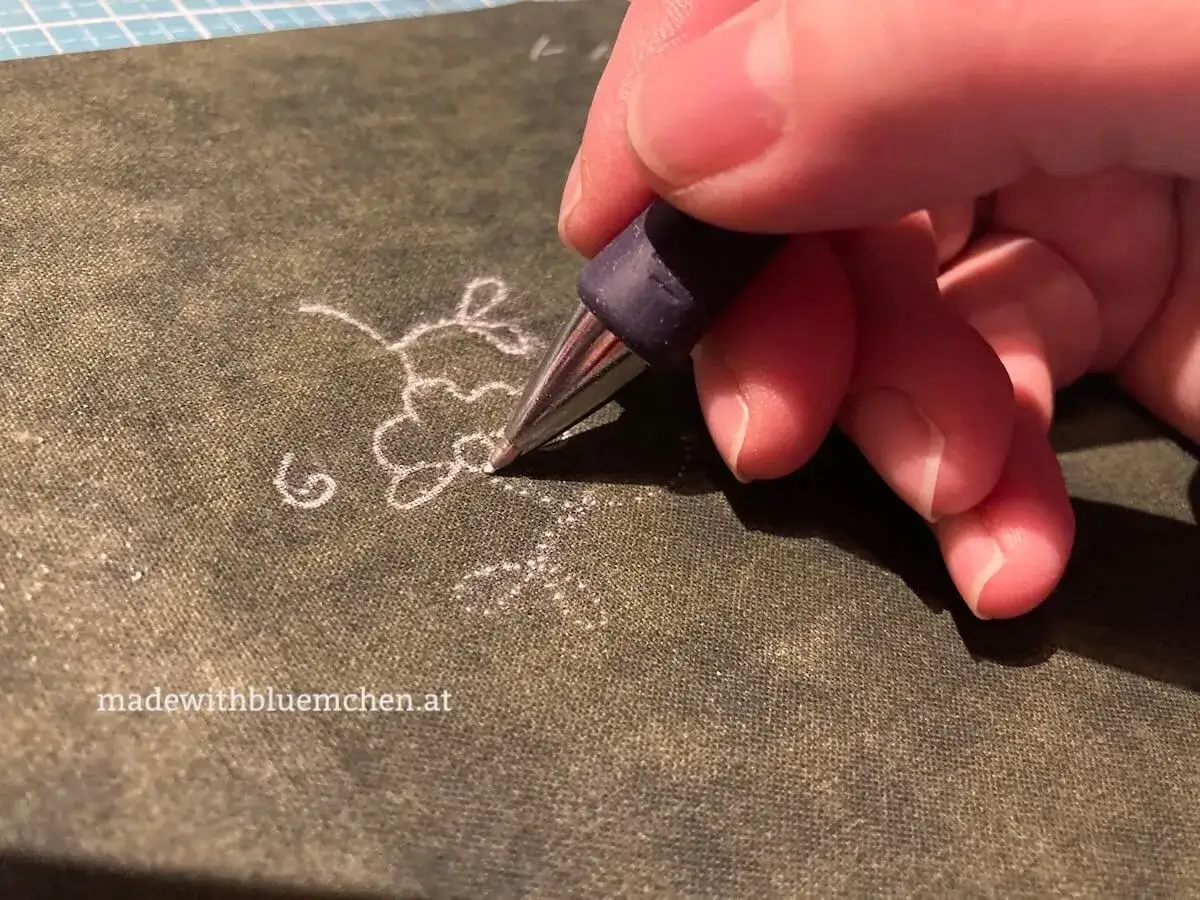 Hand &uuml;bertr&auml;gt ein zartes Blumenmuster auf Stoff. Das Stickmuster wird mit einem Stift auf den Stoff gezeichnet, bereit f&uuml;r die Stickerei.