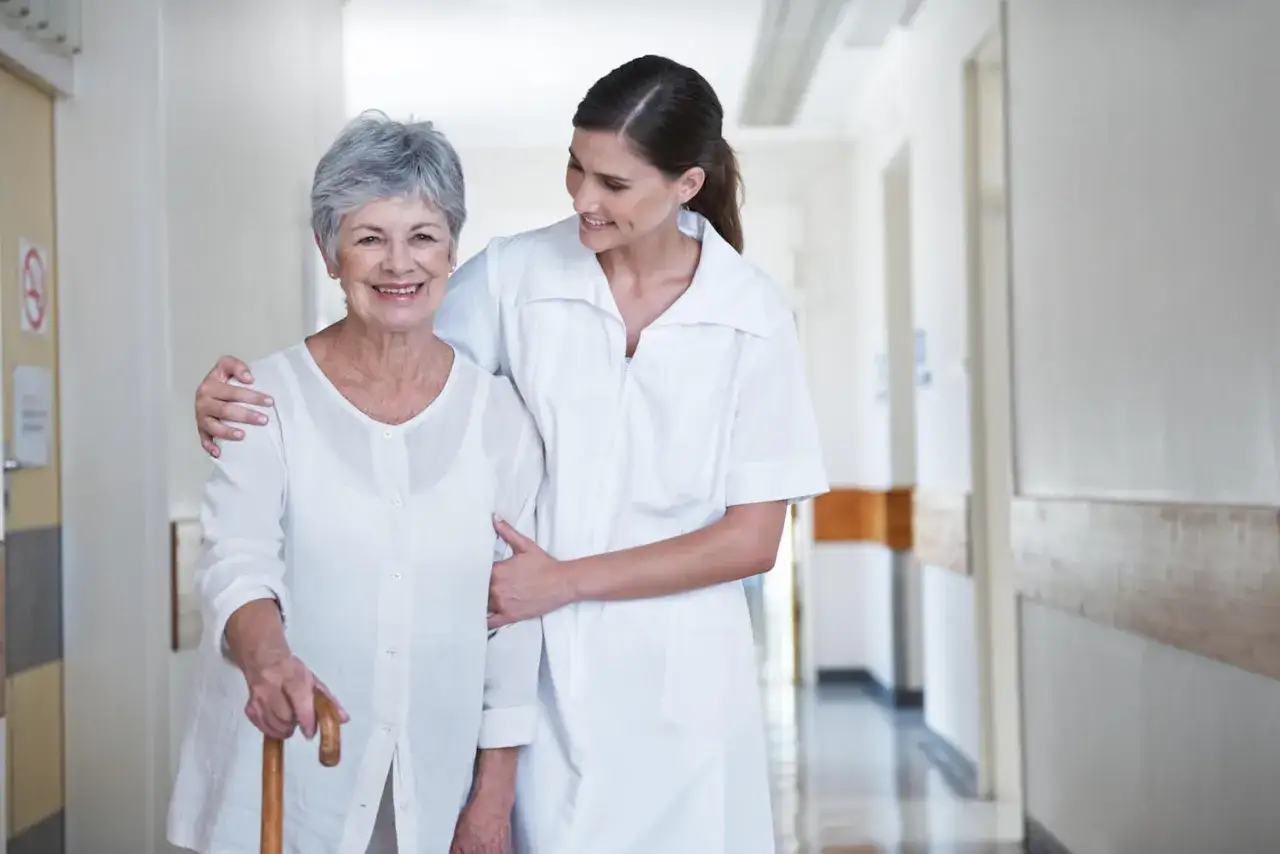 Une aide-soignante soutient une résidente souriante dans un couloir d'EHPAD. L'image évoque l'espérance de vie après entrée en ehpad, grâce à des soins attentifs.