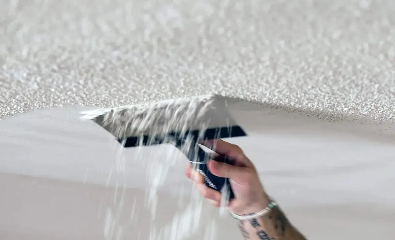 A hand scrapes away popcorn ceiling texture, sending flakes falling.