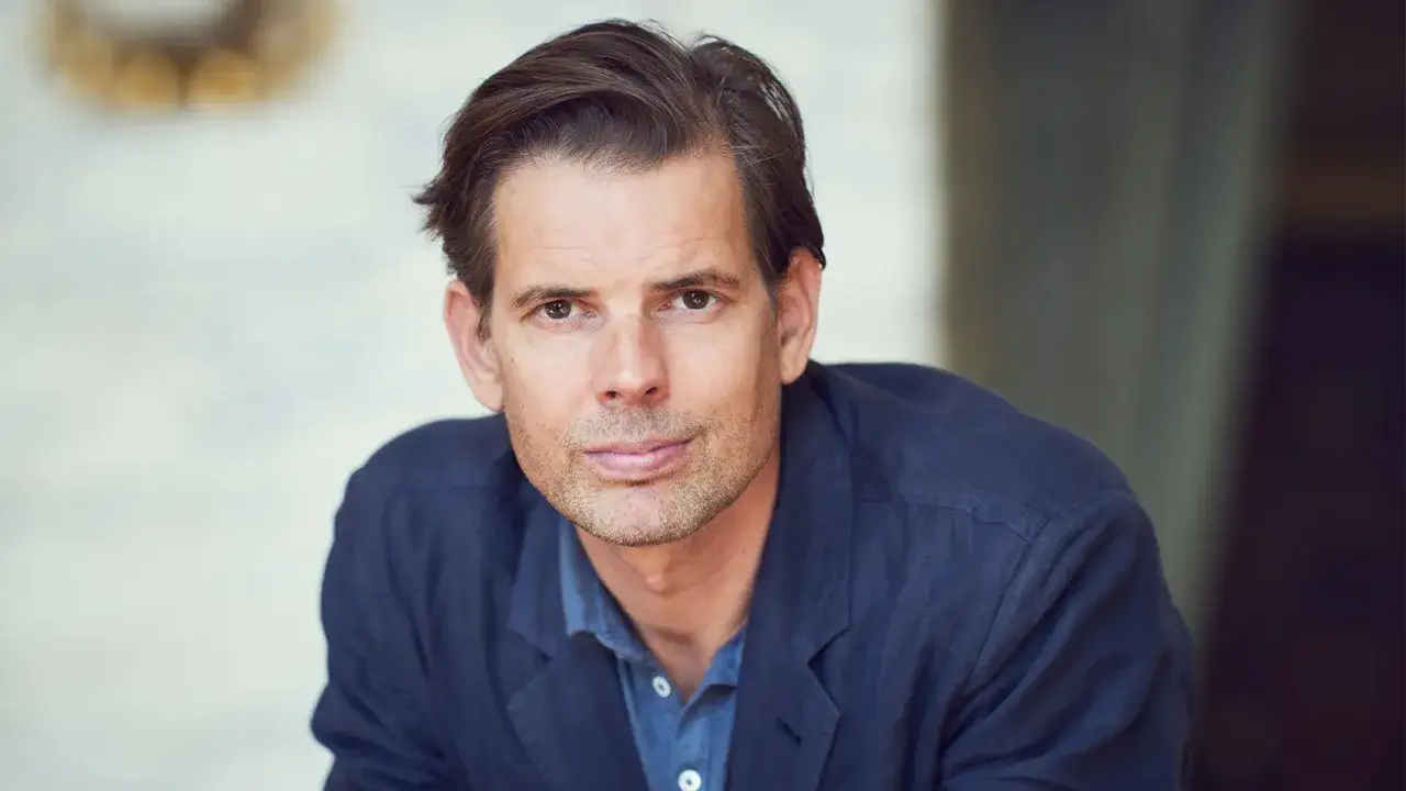 Alex Schulman, känd från Dramaten, blickar mot kameran i en mörkblå skjorta och kavaj.