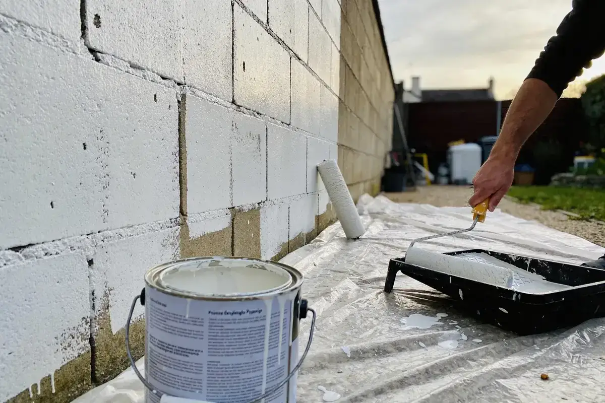 Un homme est en train de peindre un mur en parpaing avec un rouleau. La peinture blanche recouvre progressivement les blocs de béton.