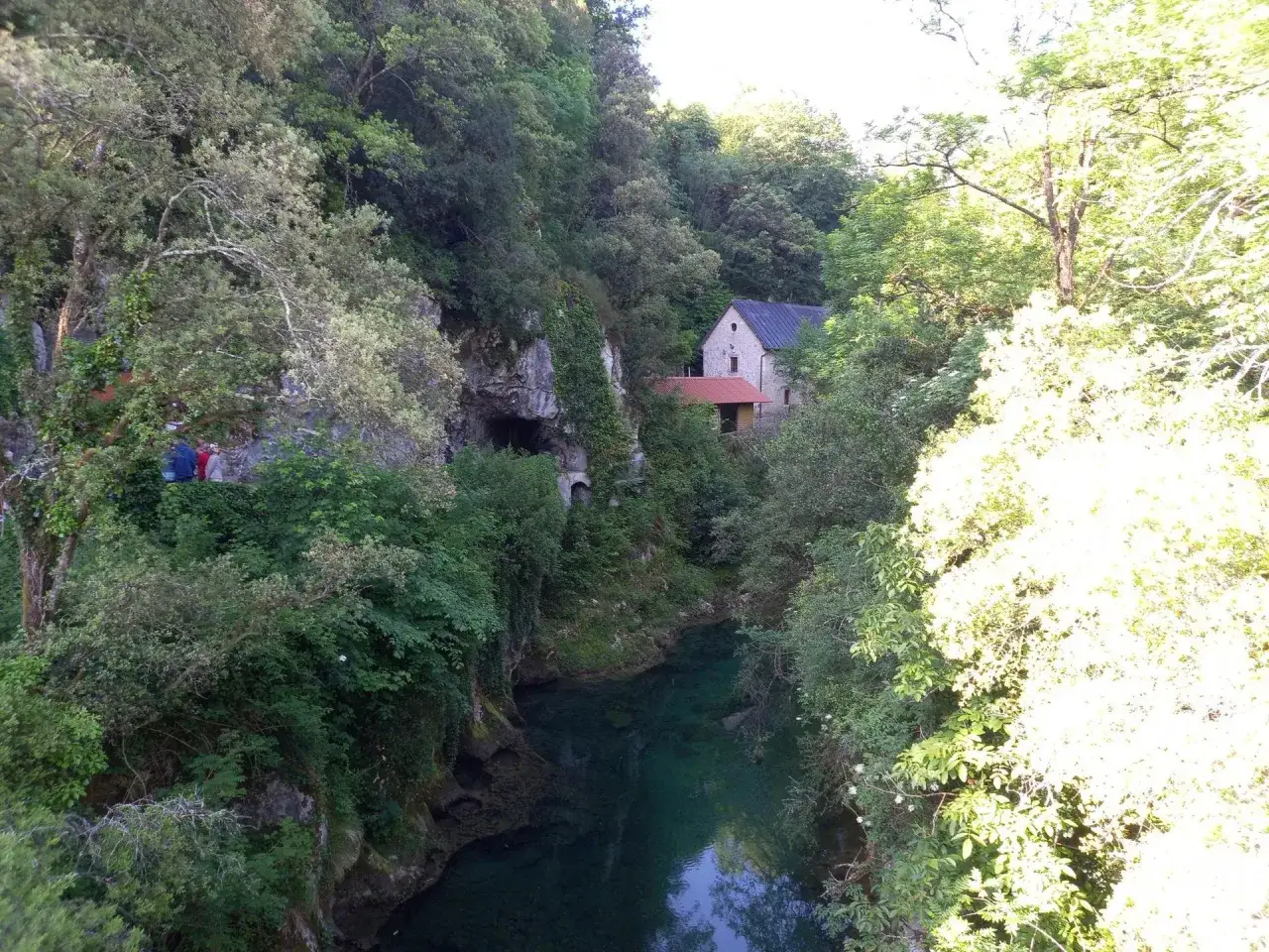Un río de aguas cristalinas fluye entre verdes bosques, cerca de las cuevas de Cangas de Onís. Una casa de piedra se alza en la ladera.