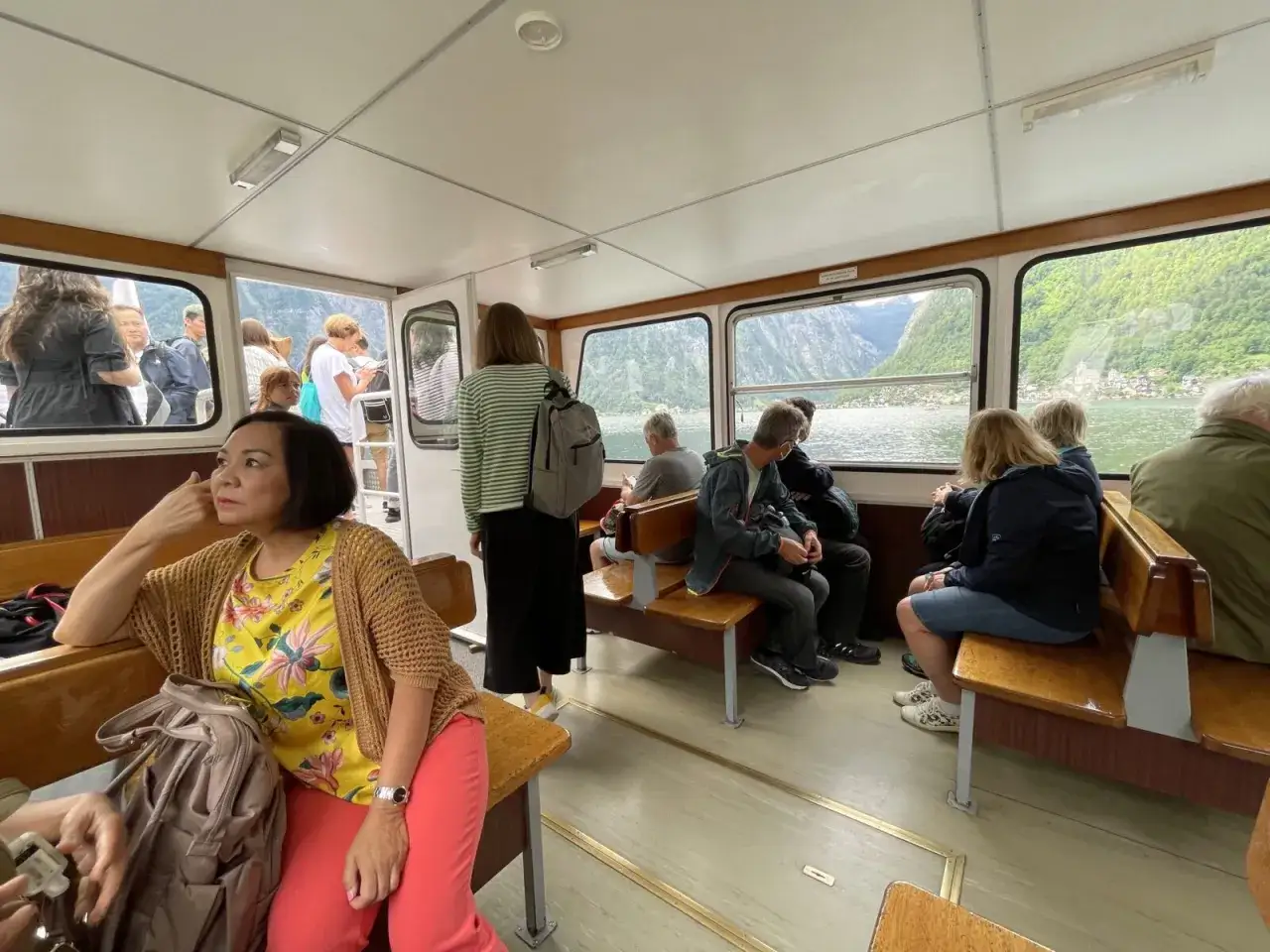 Passengers on a ferry, enjoying the scenic journey from Prague to Hallstatt. The boat glides across a lake, with lush green mountains and a charming village visible through the windows.