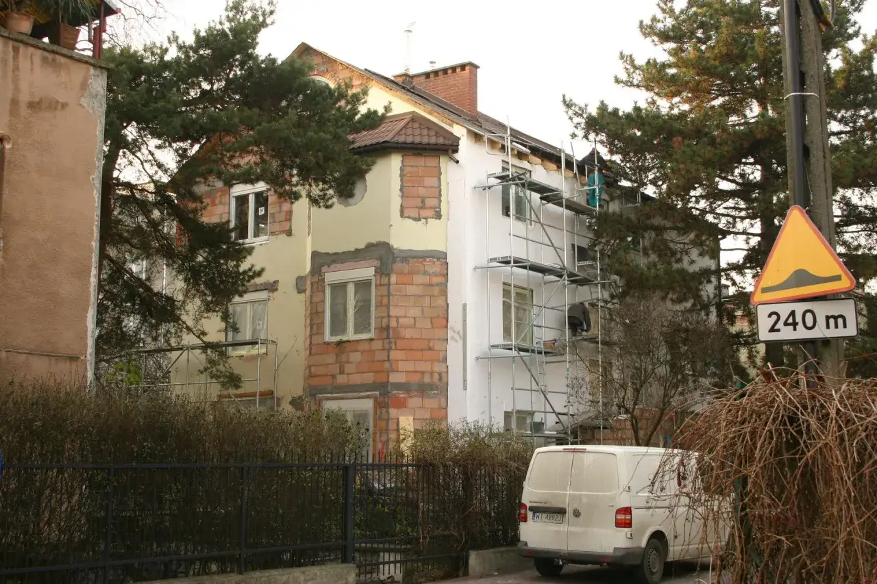 Trwa remont domu. Widać rusztowania, styropian i cegły. Obok stoi biały van.