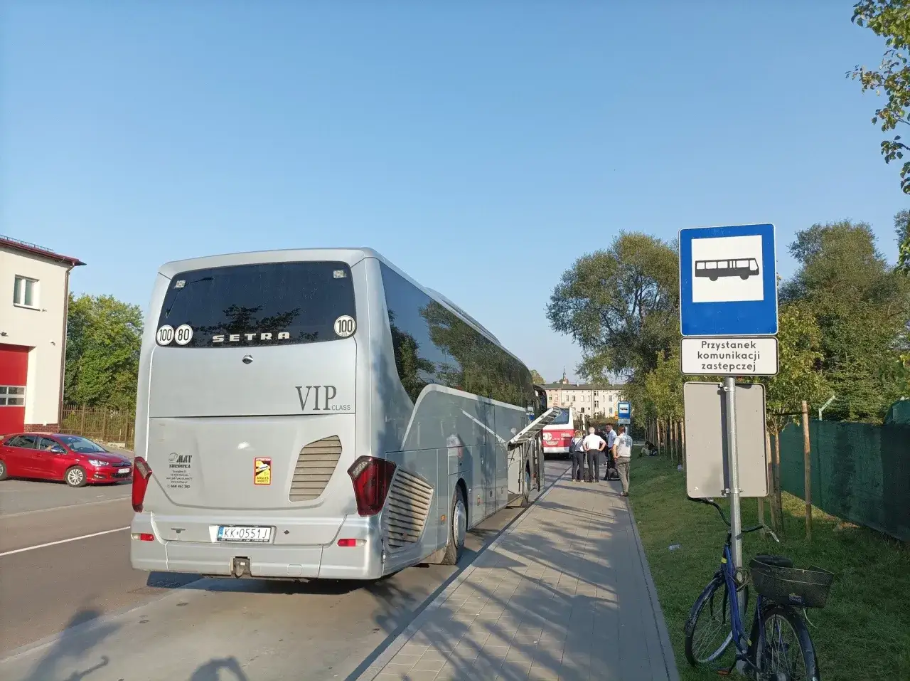 Autobus Setra VIP CLASS czeka na pasażerów na przystanku komunikacji zastępczej PKP. Obok stoi czerwony samochód.