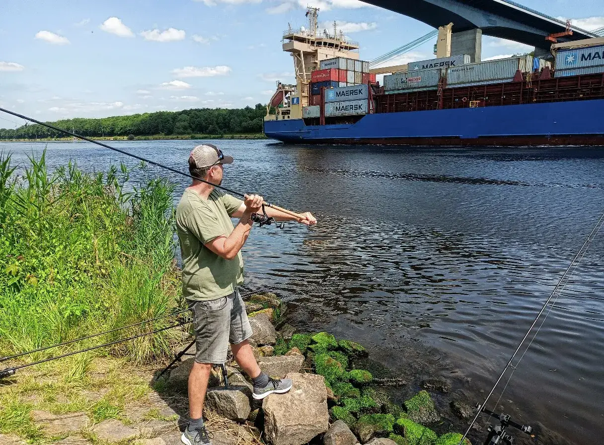 Mann mit Angelrute am Ufer, während ein Containerschiff vorbeifährt. John Sidley Rig im Einsatz.